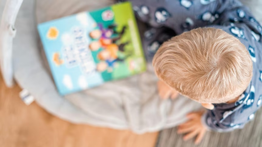 A child's head, viewed from above. They are looking at the cover of a bright children's picture book